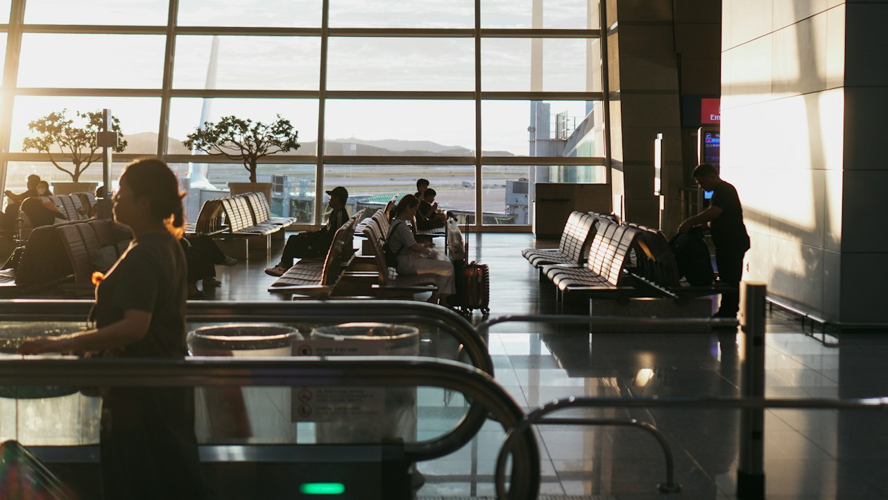 Travellers waiting with luggage at a departure gate, backlit by the morning light